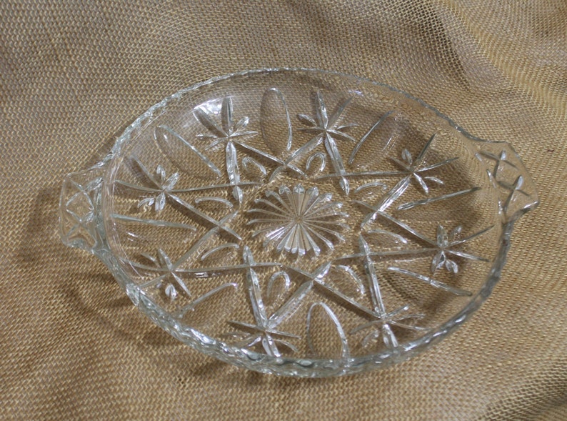 May include: A clear glass serving dish with a starburst pattern and two handles. The dish is on a brown burlap background.