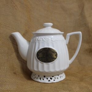 May include: White ceramic teapot with a gold label that says "TEA" and a decorative lace design on the base.