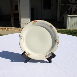 May include: A cream-colored, round plate with a gold rim and small floral clusters in orange, blue, and yellow. The plate is displayed on a black stand, set against a white tablecloth. The plate is a vintage style.