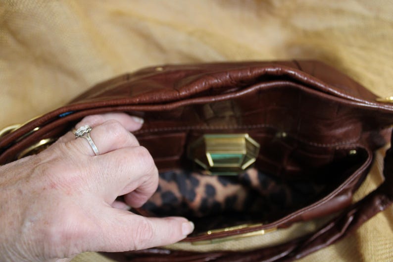 Op de afbeelding: Een bruine leren handtas met een krokodillenleertextuur. De binnenkant heeft een luipaardprint voering en een goudkleurige sluiting. De tas is open en onthult de binnenkant.
