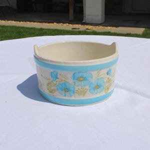 May include: A ceramic bowl with a cream-colored interior and a light blue exterior band. The bowl is decorated with blue floral patterns and has two small handles. The bowl is sitting on a white surface.