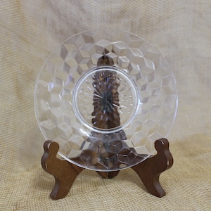 May include: A clear glass plate with a diamond pattern sits on a wooden display stand. The plate is perfect for serving desserts or appetizers.