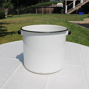 May include: A large, cylindrical white enamel stockpot with a smooth finish. The pot features a thin black rim around the top edge and two small, black side handles attached to the exterior. The interior of the pot is also white.