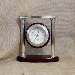 May include: A quartz mantel clock featuring a circular face with Roman numerals, suspended between two polished silver-tone metal pillars. The clock has a dark wood-toned base and a matching dark wood-toned circular frame around the clock face.