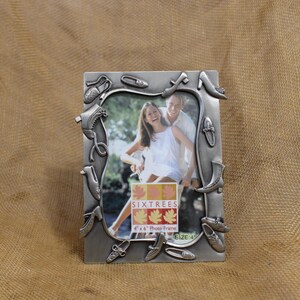 May include: A silver-toned picture frame with a design of shoes around the border. The frame is for a 4x6 inch photo and has the text "Six Trees 4" x 6" Photo Frame Size 4" printed on the bottom.