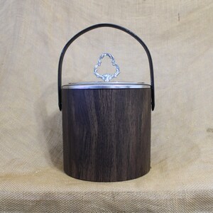 May include: A wooden ice bucket with a black leather handle and a silver lid with a decorative design. The bucket is on a brown burlap surface.