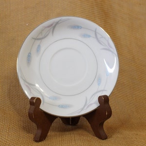 May include: A white saucer with a gold rim and a delicate wheat pattern in blue, gray, and pink. The saucer is displayed on a small, dark brown wooden stand, showcasing its design and details. This elegant piece is suitable for a tea set.