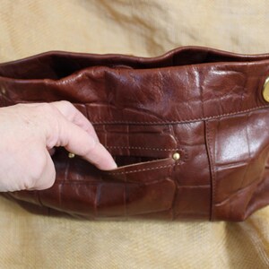 Op de afbeelding: Een bruine leren handtas met een krokodillenpatroon. De tas heeft goudkleurige hardware en een klein vakje aan de voorkant. De tas is open en onthult de binnenkant.