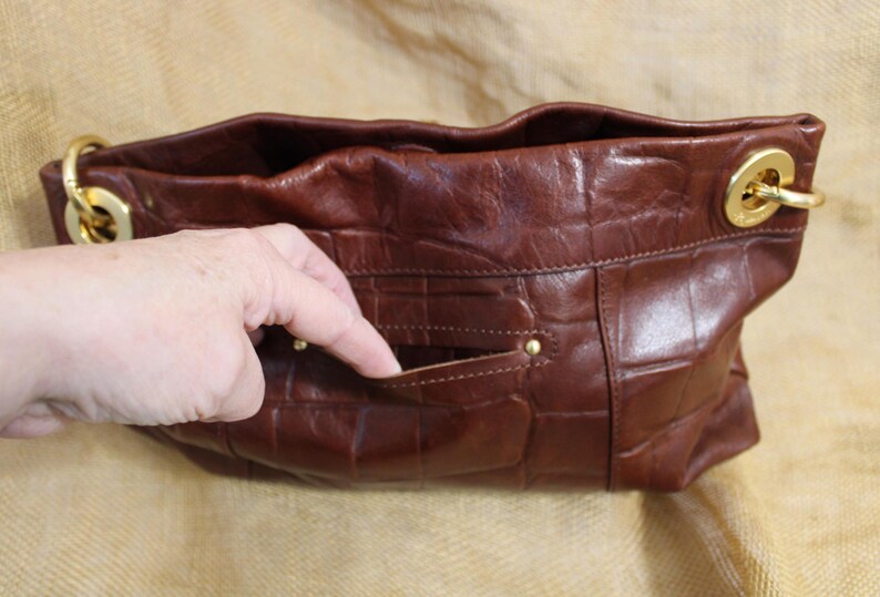 Op de afbeelding: Een bruine leren handtas met een krokodillenpatroon. De tas heeft goudkleurige hardware en een klein vakje aan de voorkant. De tas is open en onthult de binnenkant.