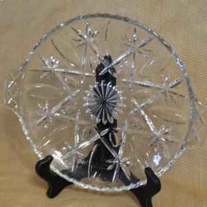 May include: A clear glass serving dish with a scalloped edge and a starburst pattern. The dish has two handles on the sides.