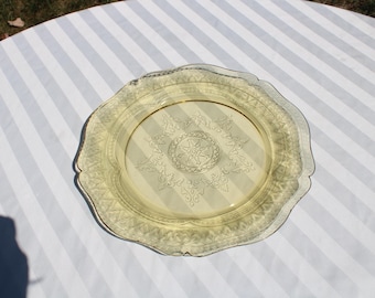 May include: A vintage yellow glass plate with a scalloped edge and intricate embossed floral and starburst design. The plate has a gold rim and is displayed on a white and gray striped surface.