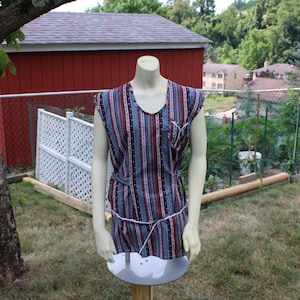 May include: A sleeveless dress with a striped pattern in shades of blue, red, and white. The dress has a V-neck and a white trim around the neckline. The dress is on a white mannequin.