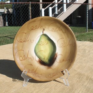 May include: A decorative ceramic plate with a hand-painted green pear and the word "Teem" repeated around the edge. The plate is on a stand.