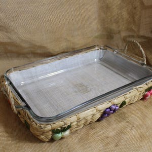 May include: A rectangular glass baking dish with a woven basket-style base. The basket is adorned with colorful floral accents. The dish has a handle made of woven material.