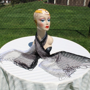 May include: A black and white ombre knit scarf with fringe on a white table. The scarf is made of a delicate, open weave material.