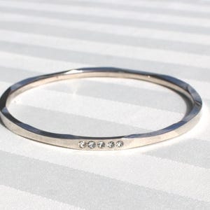 May include: A silver-toned bangle bracelet with a faceted design. The bracelet features five small, clear gemstones set in a row. The bracelet is circular and rests on a white striped surface.