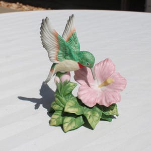 May include: A ceramic hummingbird figurine with green, white, and red details, hovering near a pink hibiscus flower. The figurine has detailed wings and is perched on a stem with green leaves. A decorative piece.