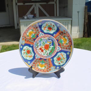 May include: Decorative ceramic plate with a colorful floral design. The plate features a blue, orange, and red color scheme with gold trim. The plate is displayed on a black stand, set against a white tablecloth.
