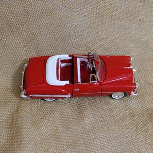 May include: A red die-cast model convertible car featuring a white interior, a white folded soft top, silver-toned trim, and white-walled tires. The vehicle is depicted from a high-angle perspective, showcasing its classic body style and open cabin.