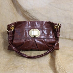 Op de afbeelding: Een bruine leren handtas met een textuur, krokodilachtig patroon. De tas heeft een goudkleurige sluiting en hardware, en een bijpassende handgreep. De tas is een middelgroot formaat en lijkt nieuw.