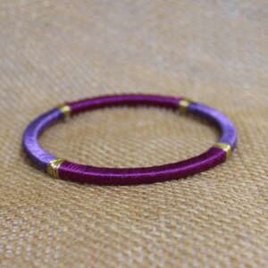 May include: A handmade bracelet with a vibrant design. The bracelet features a combination of purple and magenta threads, accented with gold-colored metal details. The bracelet is a circular shape.