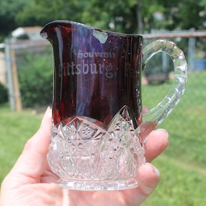 May include: A small, clear glass pitcher with a ruby red colored top. The pitcher has a cut glass design and a clear glass handle. The pitcher is inscribed with the text "Souvenir Pittsburgh, Pa."