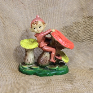 May include: A vintage ceramic figurine of a seated elf wearing a pink outfit and hat. The elf is perched on a mushroom with a red cap and yellow accents. The base is green with additional mushrooms.