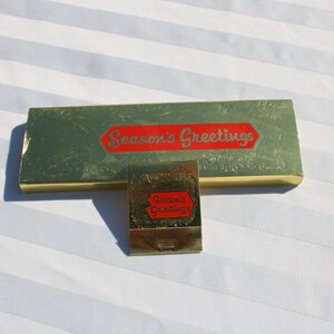 May include: A vintage gold-toned matchbox and a rectangular box, both with the text "Season's Greetings" in red. The boxes have a green background with a floral design. The items are on a white, striped surface.