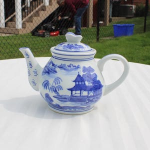 May include: A white and blue ceramic teapot with a lid. The teapot features a blue and white landscape design with a handle and spout. The teapot is sitting on a white surface.