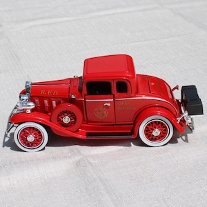 May include: A red die-cast model fire chief coupe car featuring white-walled tires, chrome headlights, and a spare tire mounted on the side. The vehicle is marked with R.F.D. on the hood and Chief on the door, with a black trunk mounted on the rear.