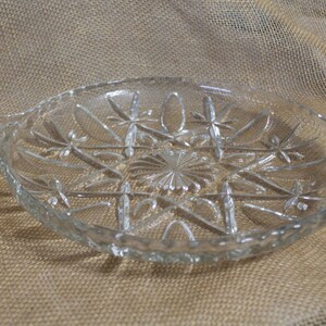 May include: A clear glass serving dish with a starburst pattern and two handles. The dish is on a brown burlap background.