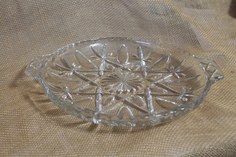 May include: A clear glass serving dish with a starburst pattern and two handles. The dish is on a brown burlap background.