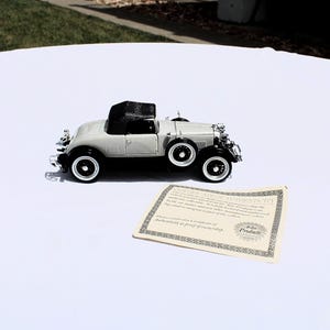 May include: A light gray and black vintage-style model car with a black convertible top, white-walled tires, and a spare wheel mounted on the side. Beside the car is a rectangular certificate of authenticity with black text and a decorative border.