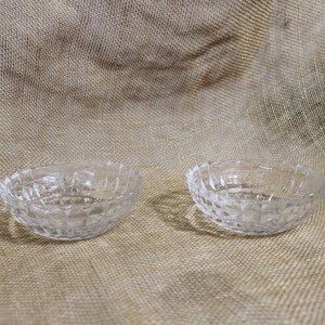 May include: Two clear, small, glass bowls with a diamond pattern. The bowls are oval-shaped and sit on a textured, tan surface. The bowls are empty and appear to be decorative or for serving small portions.