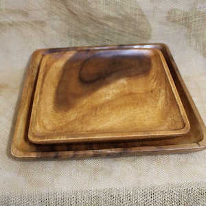 May include: Two square wooden serving trays, one nested inside the other. The trays are made of light brown wood with a natural grain pattern.