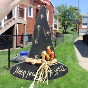 May include: A black wooden witch hat decoration with gold stars and the text "Hang Around A Spell". The hat features a straw brim and candy corn accents, perfect for Halloween or fall decor.