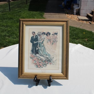 May include: A framed vintage illustration of a wedding scene. The bride and groom are in the foreground, with three bridesmaids behind them. The bride is wearing a white dress and veil, and the groom is wearing a black suit. The illustration is in a gold frame and is on a white table.