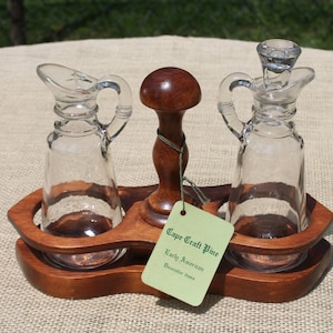 May include: Wooden oil and vinegar cruet set with two clear glass bottles and a wooden base. The bottles have a spout and a handle. The base has a label that reads "Cape Craft Pine Early American Breadboard Style".