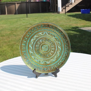 May include: A decorative plate with a teal and gold concentric design. The plate is supported by a black stand and sits on a white striped surface. The plate's intricate pattern is visible.