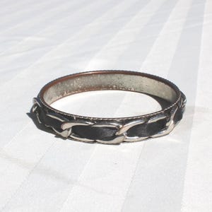 Puede incluir: Un brazalete plateado con una banda negra y un diseño de eslabones de cadena. El brazalete tiene forma circular y está sobre un fondo blanco a rayas. Un accesorio de moda.