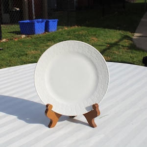 May include: A white ceramic plate with a raised pattern around the edge is displayed on a wooden stand. The plate is on a white tablecloth with stripes.