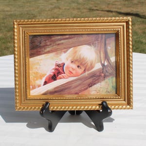 May include: A framed painting featuring a young child with blonde hair, resting on a wooden fence. The child wears a red plaid shirt. The gold frame has a rope-like design. The artwork is displayed on a black easel.
