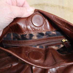 Op de afbeelding: Een bruine leren handtas met een luipaardprint voering. De tas heeft een magnetische druksluiting en een gouden rits. De binnenkant is zichtbaar, met de voering en een klein vakje.