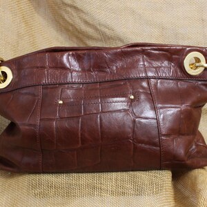 Op de afbeelding: Een bruine leren handtas met een krokodillenpatroon. De tas heeft goudkleurige hardware, waaronder ringen en kleine studs. De tas heeft een diepe, rijke bruine kleur.