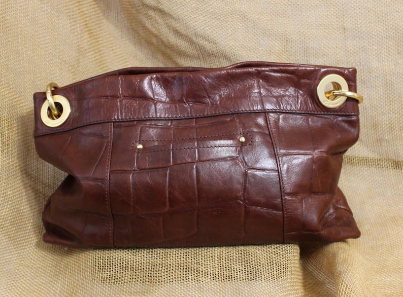 Op de afbeelding: Een bruine leren handtas met een krokodillenpatroon. De tas heeft goudkleurige hardware, waaronder ringen en kleine studs. De tas heeft een diepe, rijke bruine kleur.