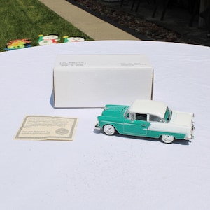 May include: A teal and white classic car model with a white roof, parked on a white surface. A white box and a certificate are also on the surface. The car model is a vintage style.