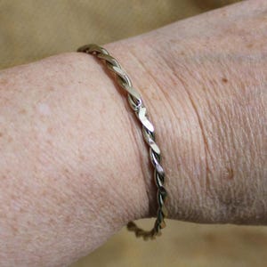May include: A silver-toned, twisted metal bracelet. The bracelet is worn on a wrist, and the design features a braided or rope-like pattern. The metal has a shiny, reflective surface, and the overall style is simple and elegant.