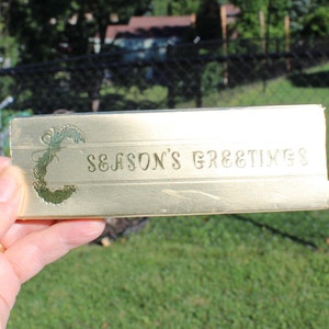 May include: A rectangular, gold-colored card with the words "SEASON'S GREETINGS" embossed on the front. The card has a decorative floral design on the left side. The card is being held in front of a green background.