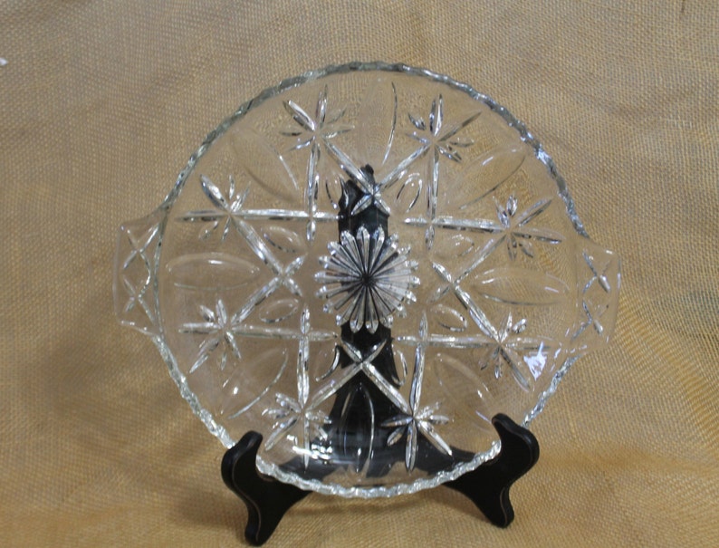 May include: A clear glass serving dish with a scalloped edge and a starburst pattern. The dish has two handles on the sides.
