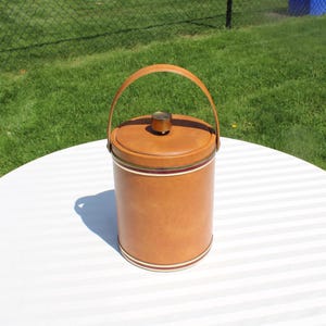 May include: A vintage brown ice bucket with a matching lid and handle. The cylindrical bucket has a decorative band of white, red, and green near the base. The handle is made of the same material as the bucket. The ice bucket is sitting on a white table.